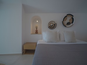 A bedroom with a bed, a bedside table, a wall-mounted shelf with a vase, and two wall decorations. - Anemos Family Suites (Santorini)