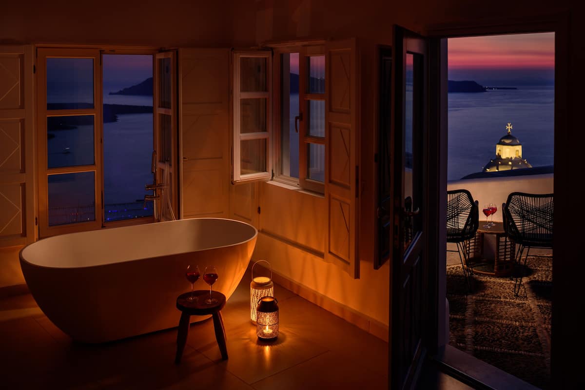 A room with a bathtub, a small table with a wine glass, and a view of a sunset over the sea.