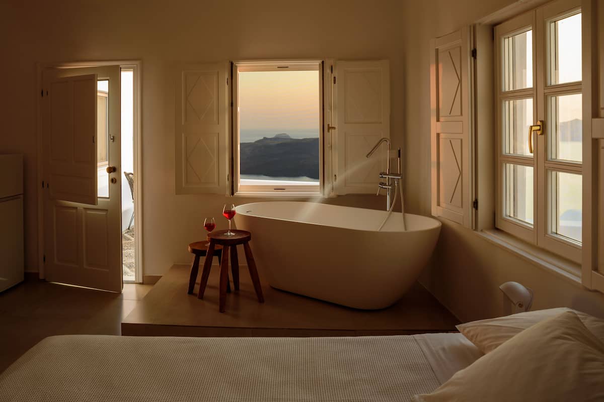 A modern bathroom with a freestanding bathtub, a window with a view of mountains, and a small wooden stool with a glass of wine.