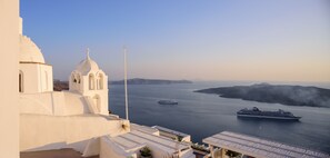 A white building with a dome and a cross, overlooking a calm sea with a cruise ship and a smaller boat. - Anemos Family Suites (Santorini)