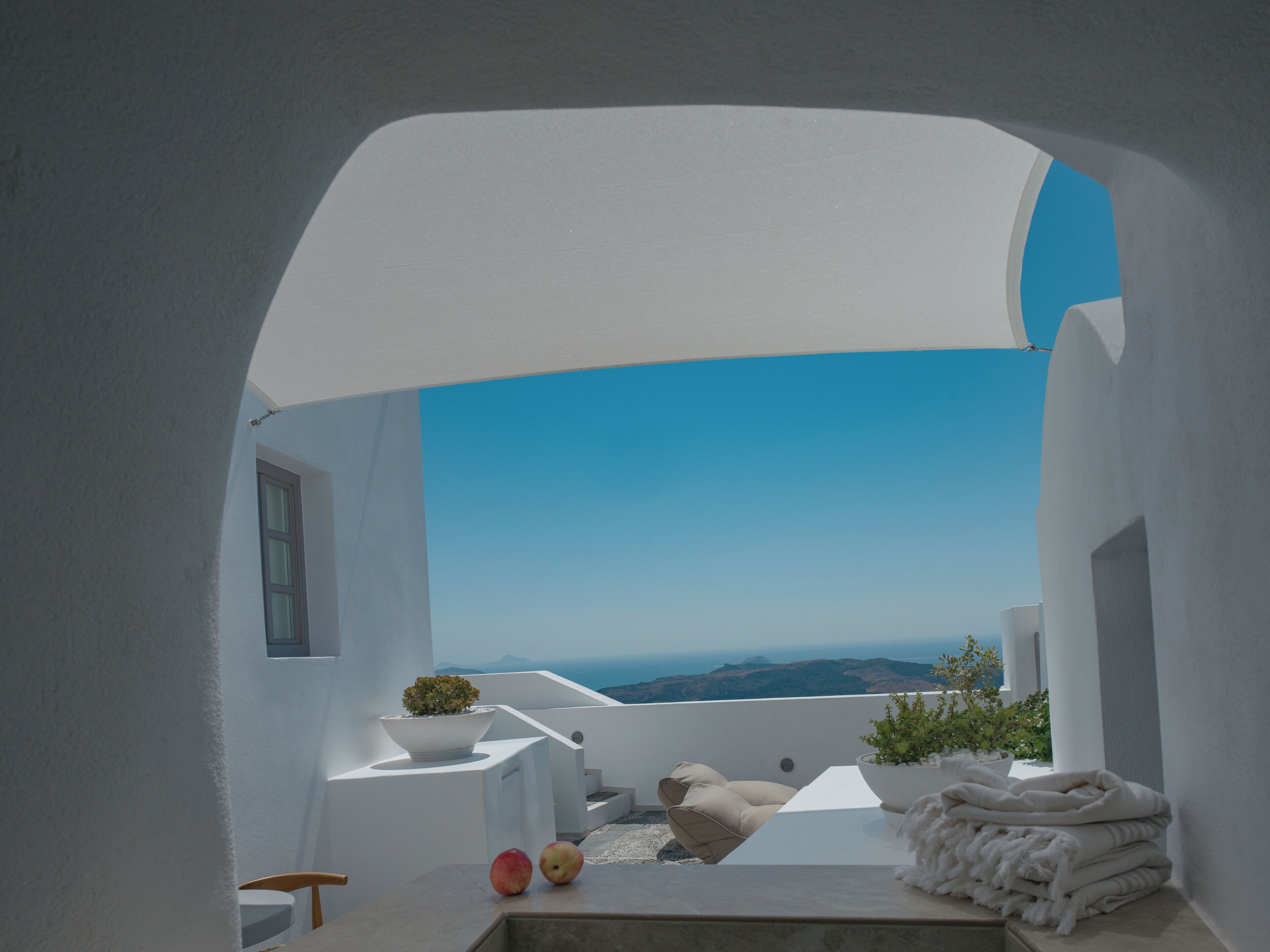 Um espaço ao ar livre moderno e minimalista, com vista clara para o mar, uma mesa branca com uma planta e uma pilha de toalhas.