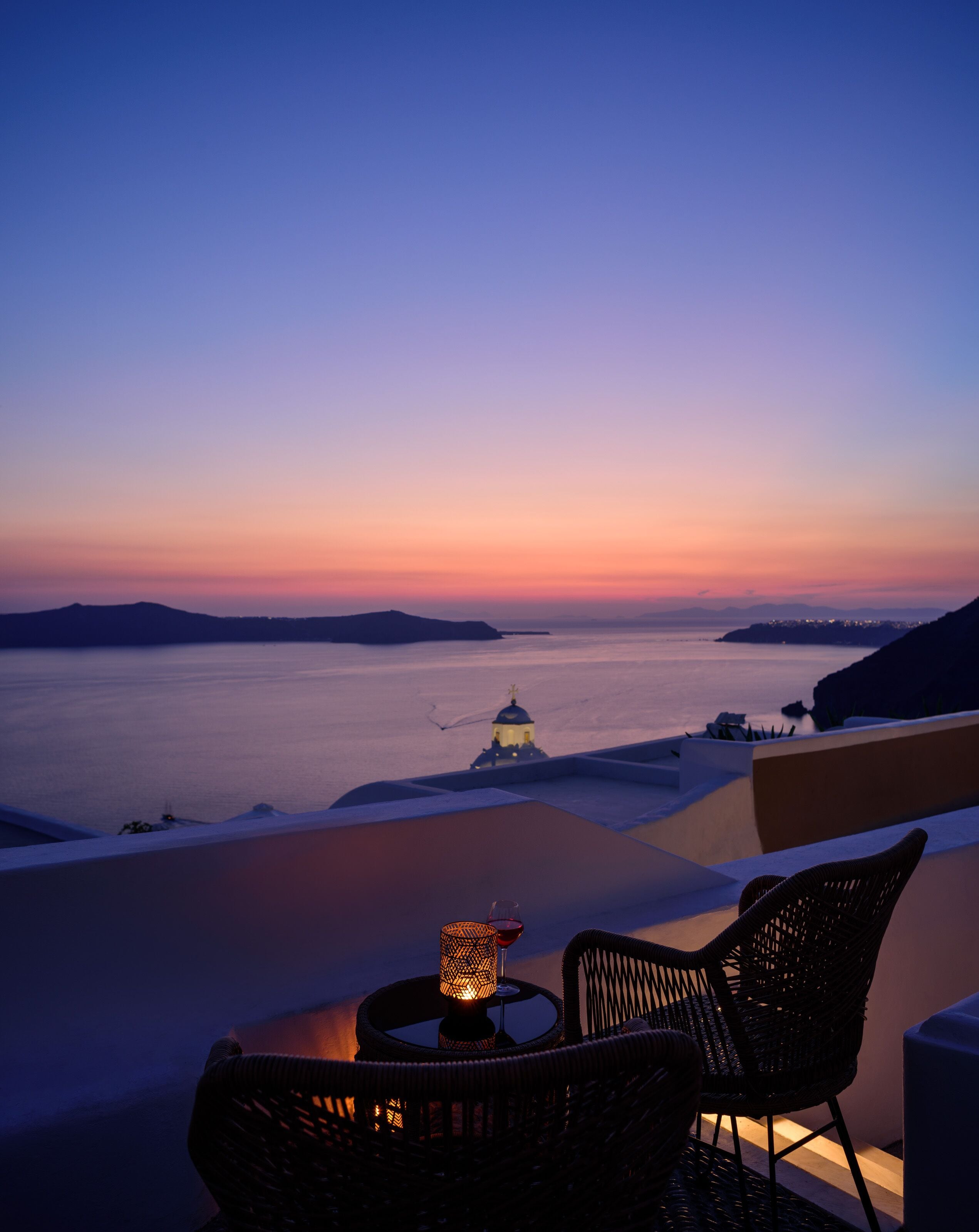 A sunset view from a balcony with wicker chairs and a table, overlooking a calm sea and distant islands.