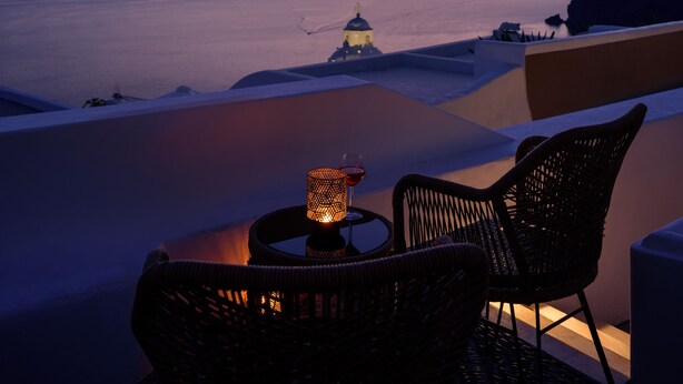 A sunset view from a balcony with wicker chairs and a table, overlooking a calm sea and distant islands. - Anemos Family Suites (Santorini)