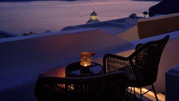 A sunset view from a balcony with wicker chairs and a table, overlooking a calm sea and distant islands.