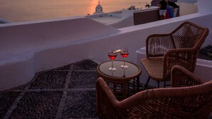 A sunset view from a terrace with wicker chairs and a table with glasses of red wine. - Anemos Family Suites (Santorini)