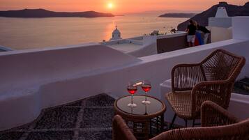 Vista al atardecer desde una terraza con sillas de mimbre y una mesa con copas de vino tinto.