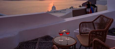 Vista al atardecer desde una terraza con sillas de mimbre y una mesa con copas de vino tinto.