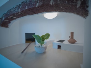 A room with a white wall, a brick arch, a potted plant, a small table with a vase, and a wall-mounted shelf. - Anemos Family Suites (Santorini)