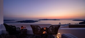 A terrace with wicker chairs and a table, overlooking a sunset view of the sea and islands. - Anemos Family Suites (Santorini)