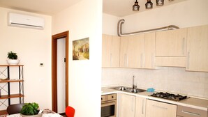 A modern kitchen with light wood cabinets, a stainless steel sink, and a gas stove. There is a dining area with a table covered by a lace tablecloth and a potted plant. - Diana apartment, near the historic center bright and spacious (Oderzo)