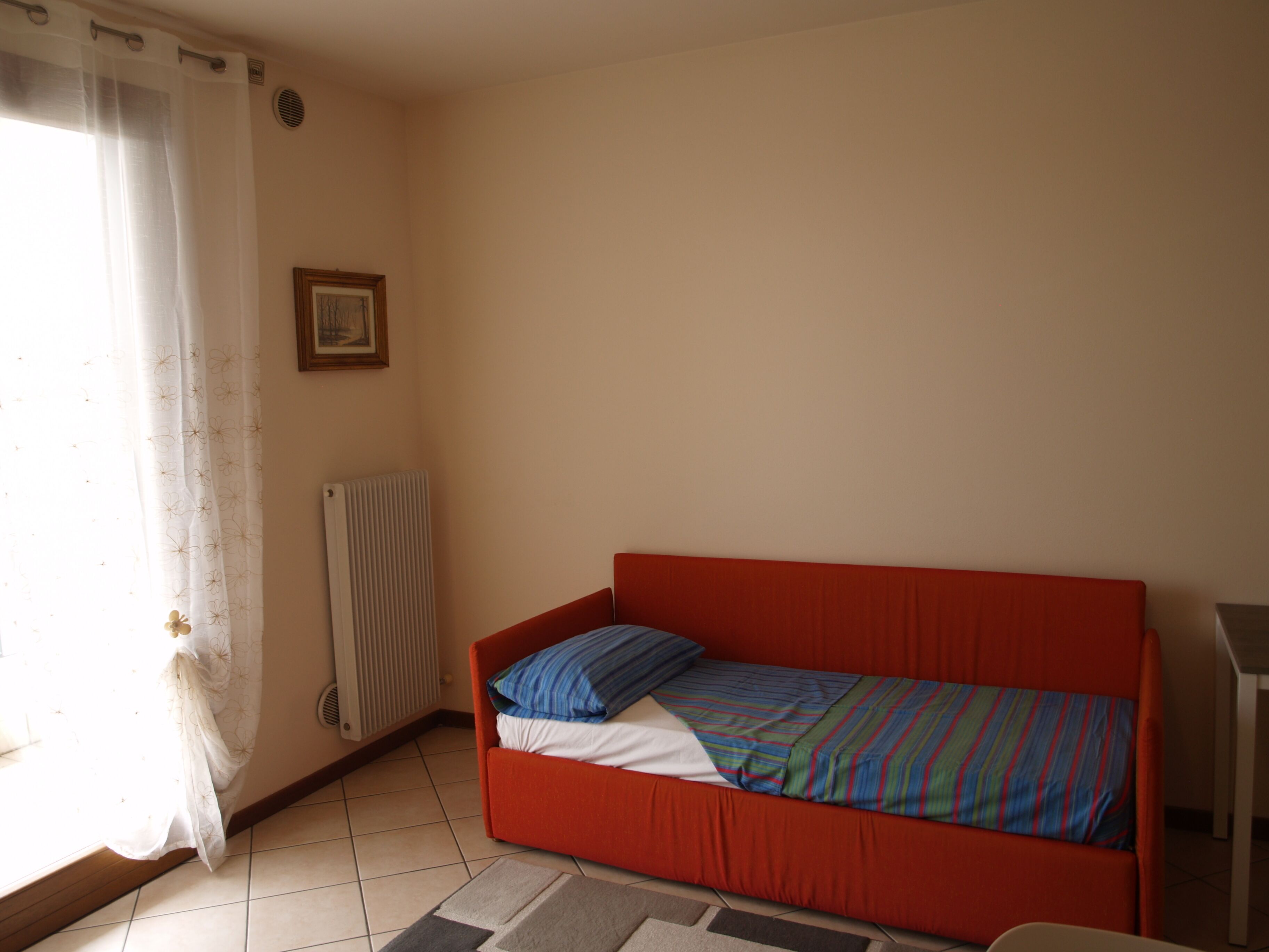 Una habitación pequeña con cama individual, una cama roja, un radiador, una ventana con cortinas traslúcidas y una mesita.