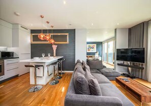 Een moderne woonkamer met een grijze bank, een houten salontafel en een televisie. Er is een keuken met een wit aanrecht en barkrukken.