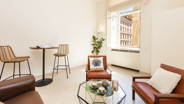 A modern living room with a glass coffee table, two wicker chairs, a brown leather sofa, and a view of a building through a large window. - Lovely Cozy Nazionale 4 (Roma)