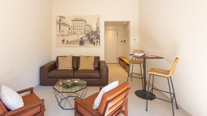 A living room with a brown leather sofa, a glass coffee table, and a wicker chair. There is a painting on the wall depicting a street scene. - Lovely Cozy Nazionale 4 (Roma)