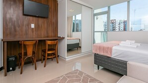 Ett modernt hotellrum med ett skrivbord i trä, två stolar, en säng med en grå sänggavel och ett stort fönster med utsikt över staden.