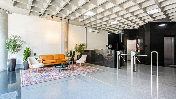 Eine moderne Lobby mit geometrischer Decke, einer Ledersofa, zwei weißen Stühlen, einem gemusterten Teppich und einer Rezeption.