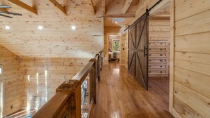 A wooden interior with a wooden staircase, a sliding barn door, and a hallway leading to a room with a large window. - Lake Lure's Newest Luxury Log Cabin Behind The Gates of Rumbling Bald Resort! (Lake Lure)