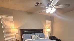 A bedroom with a bed, nightstands, a ceiling fan, and a window with blinds. - The House with the Blue Door (Norman)