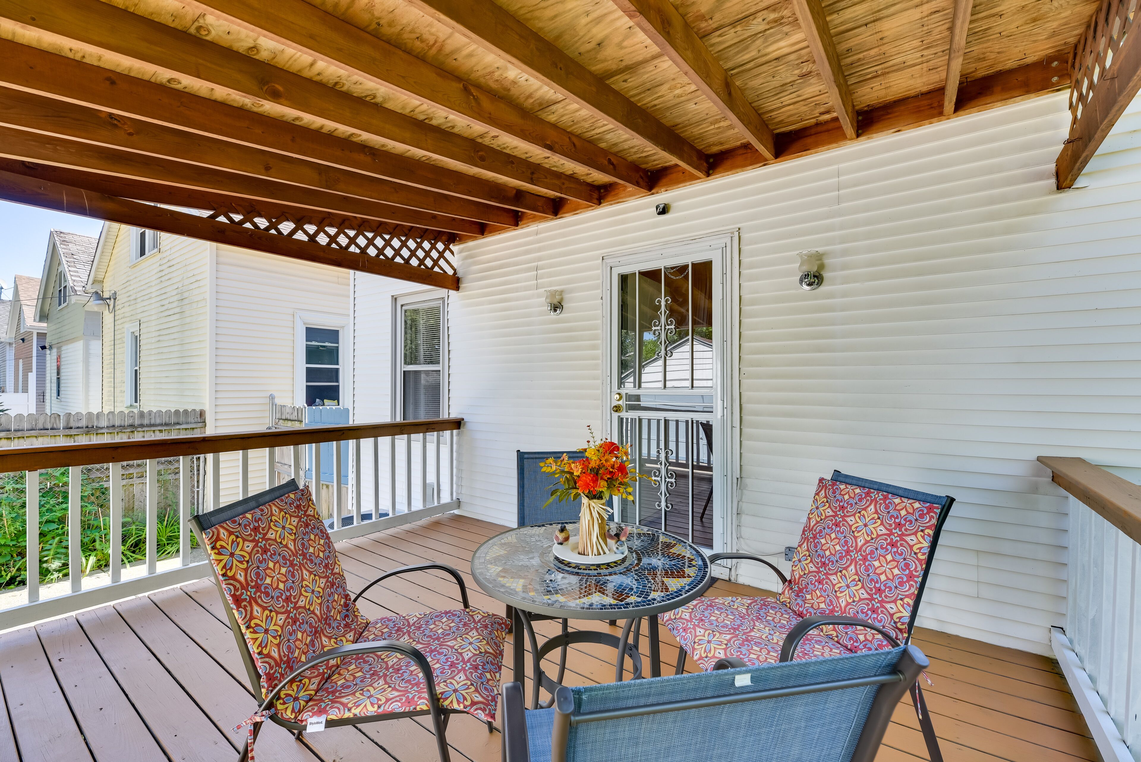 A covered porch with a wooden ceiling, a white exterior wall, and a glass table with chairs.