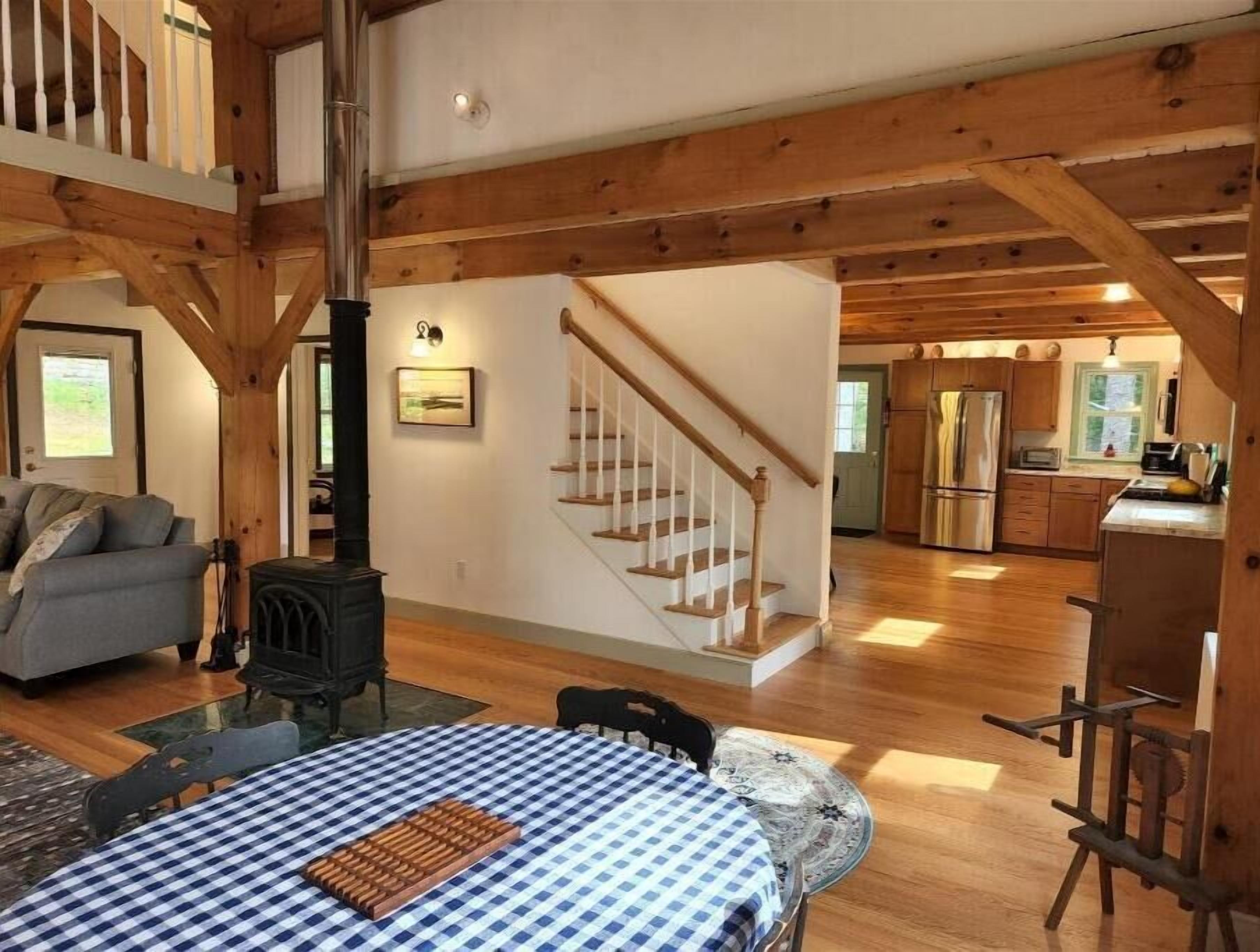 Un vaste espace de vie doté d’un plafond haut, de poutres en bois, d’une cheminée, d’une table à manger recouverte d’une nappe à carreaux et d’une cuisine située au fond de la pièce.