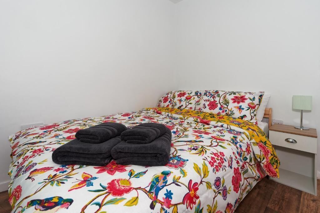 A neatly made bed with a floral duvet, a pair of folded towels, and a bedside table with a lamp.