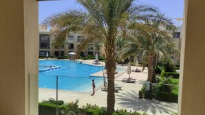 Vista de una piscina con palmeras y sillones de descanso, rodeada por un edificio de varios pisos.