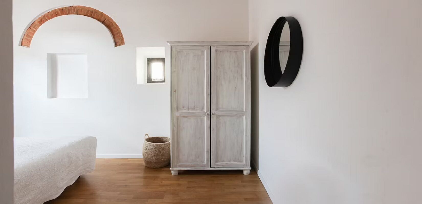 A bedroom with a bed, a wardrobe, a round mirror, and a small window.