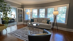 A living room with a sofa, two side tables, a coffee table, and a chair. There are large windows with blinds, a plant, and a lamp. - Spacious 2 bed room apartment near Shaker, Cleveland clinic, CWRU, Van aken.  (Cleveland Heights)