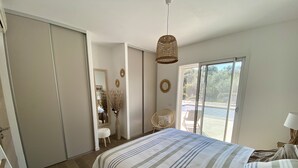 Ein Schlafzimmer mit einem großen Bett, einer Hängelampe, einem Sessel, einem Schminktisch mit Spiegel und Blick ins Freie durch eine Schiebetür.