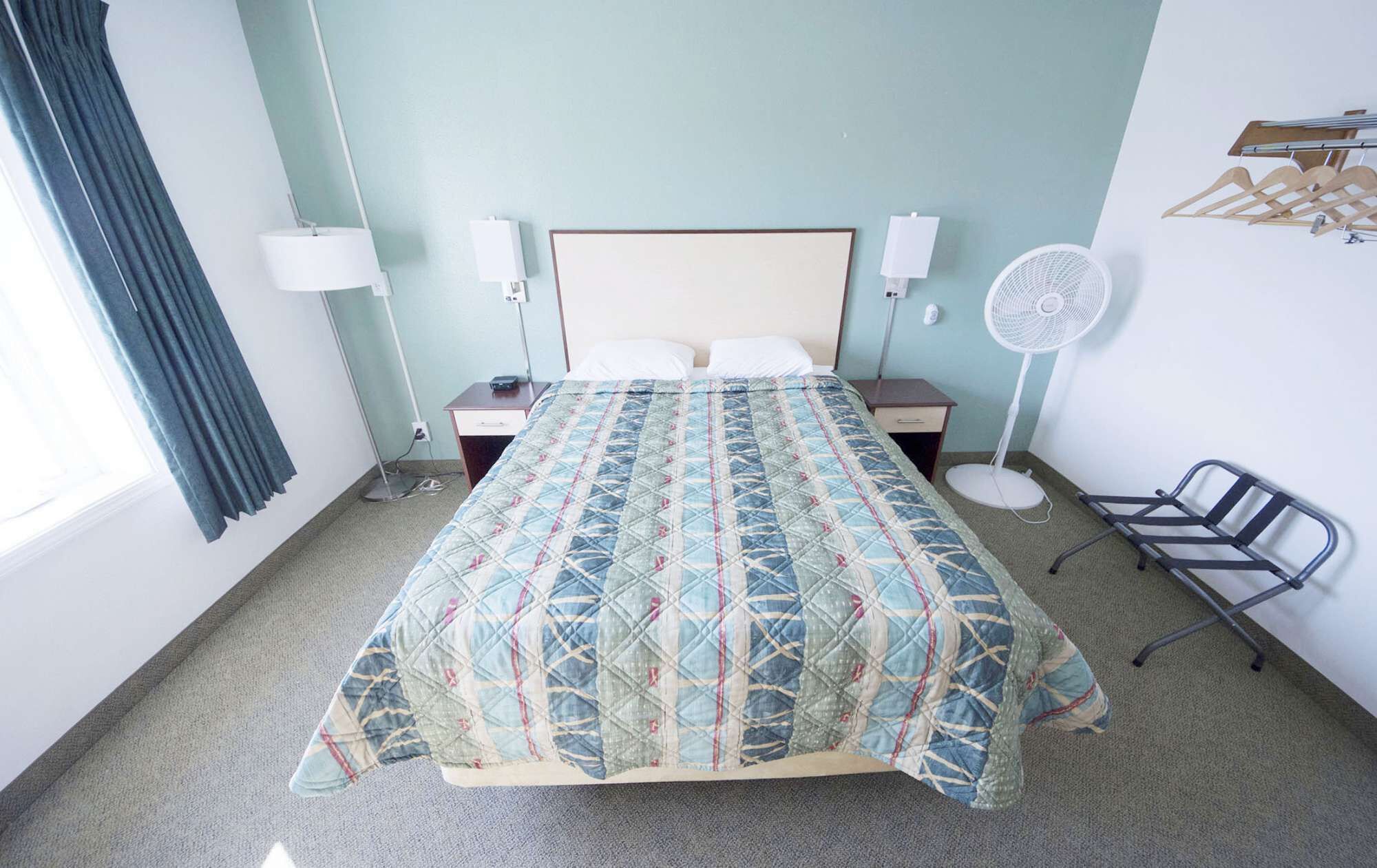 A hotel room with a bed, bedside lamps, a standing fan, a chair, and a coat rack.