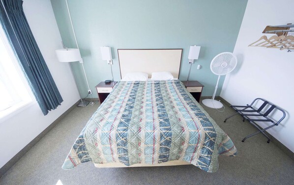 Habitación de hotel con cama, lámparas de noche, ventilador de pie, una silla y perchero.