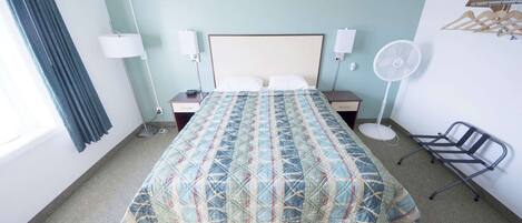A hotel room with a bed, bedside lamps, a standing fan, a chair, and a coat rack.