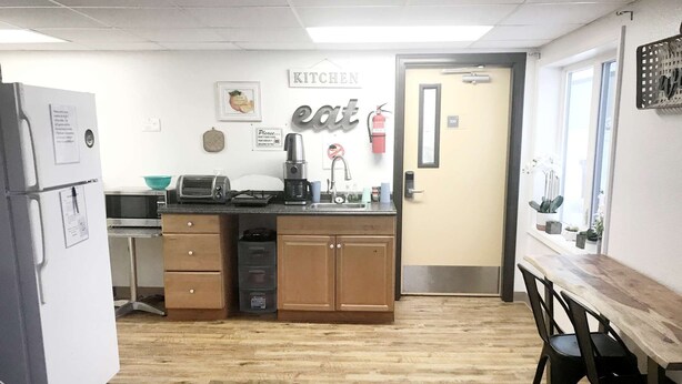 A compact kitchen with a microwave, toaster, coffee maker, and a sink. There's a door labeled '100' and a small window with plants. - Rewak Studios: Gigabit Powered WiFi! Hotel-Style Furnished Studios--Easy Walk to Lots of Places (Fairbanks)