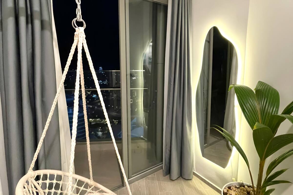 A balcony with a white macrame hanging chair, a potted plant, and a view of the cityscape at night.