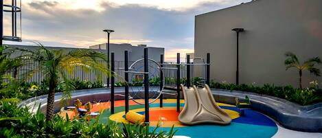 A playground with slides and climbing structures, surrounded by greenery and modern buildings.