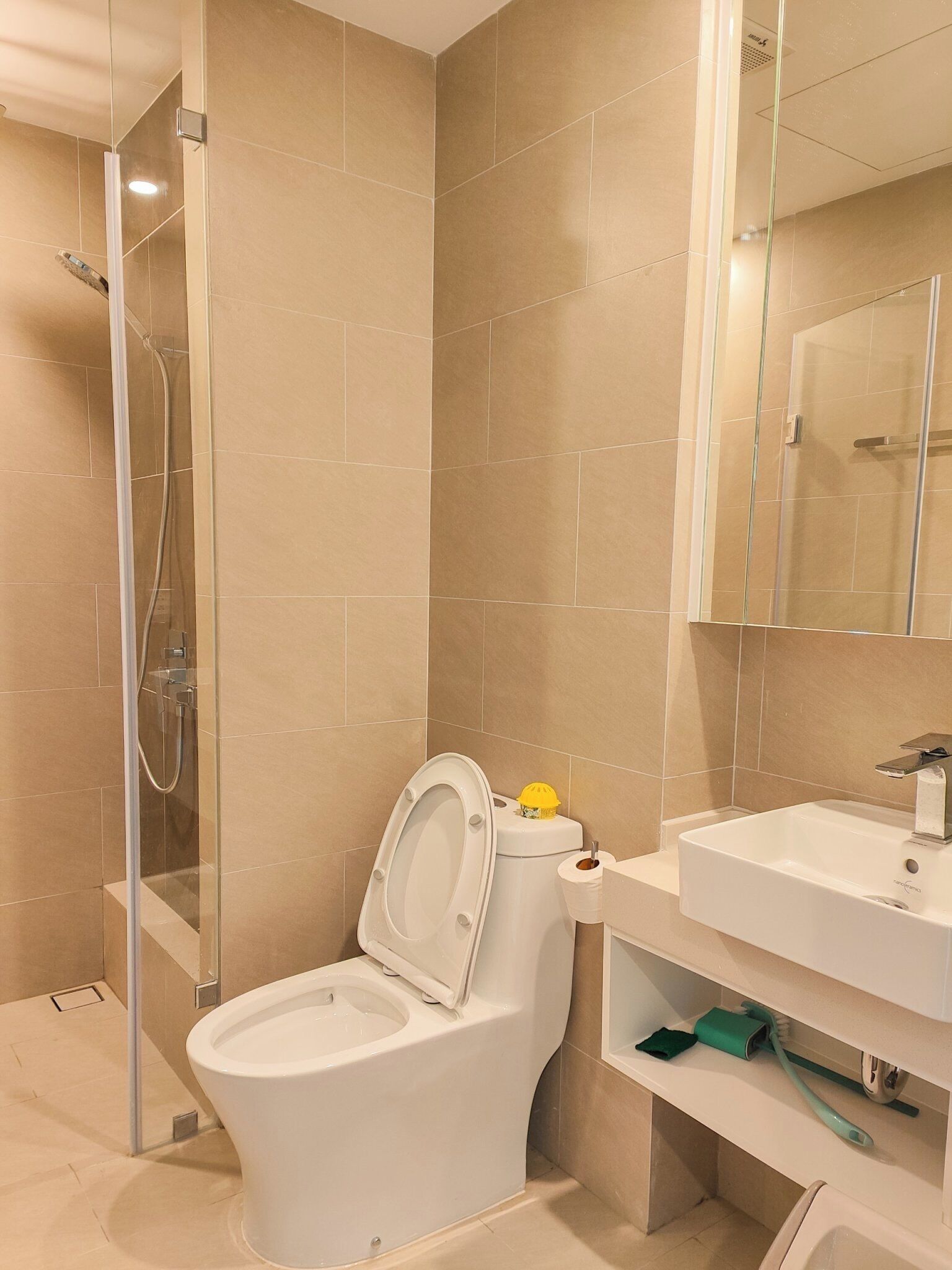 A modern bathroom with a glass-enclosed shower, a toilet with a yellow safety cap, and a sink with a mirror above it.