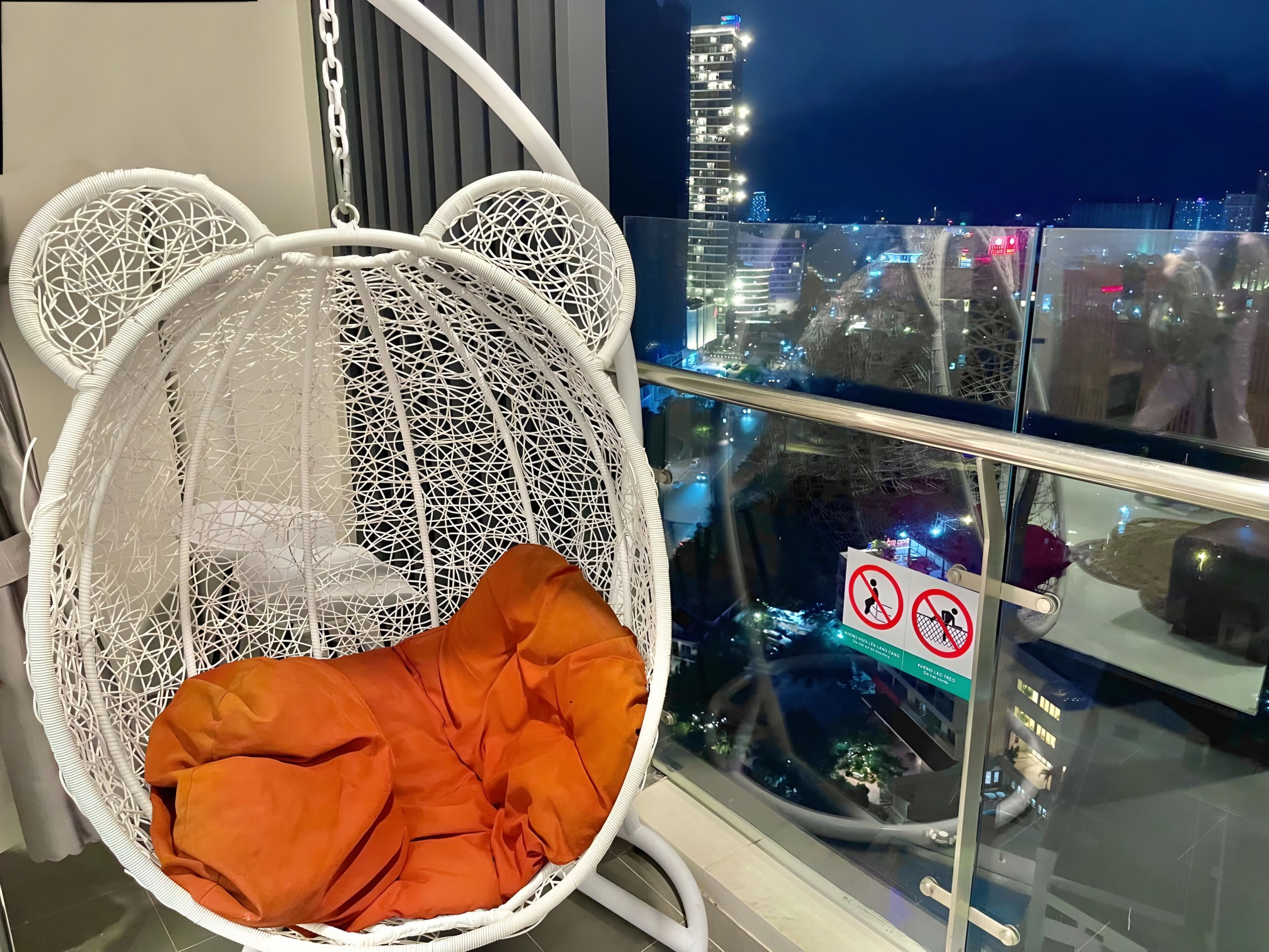 A white hanging chair with a bear design, an orange cushion, and a city view through a glass railing.