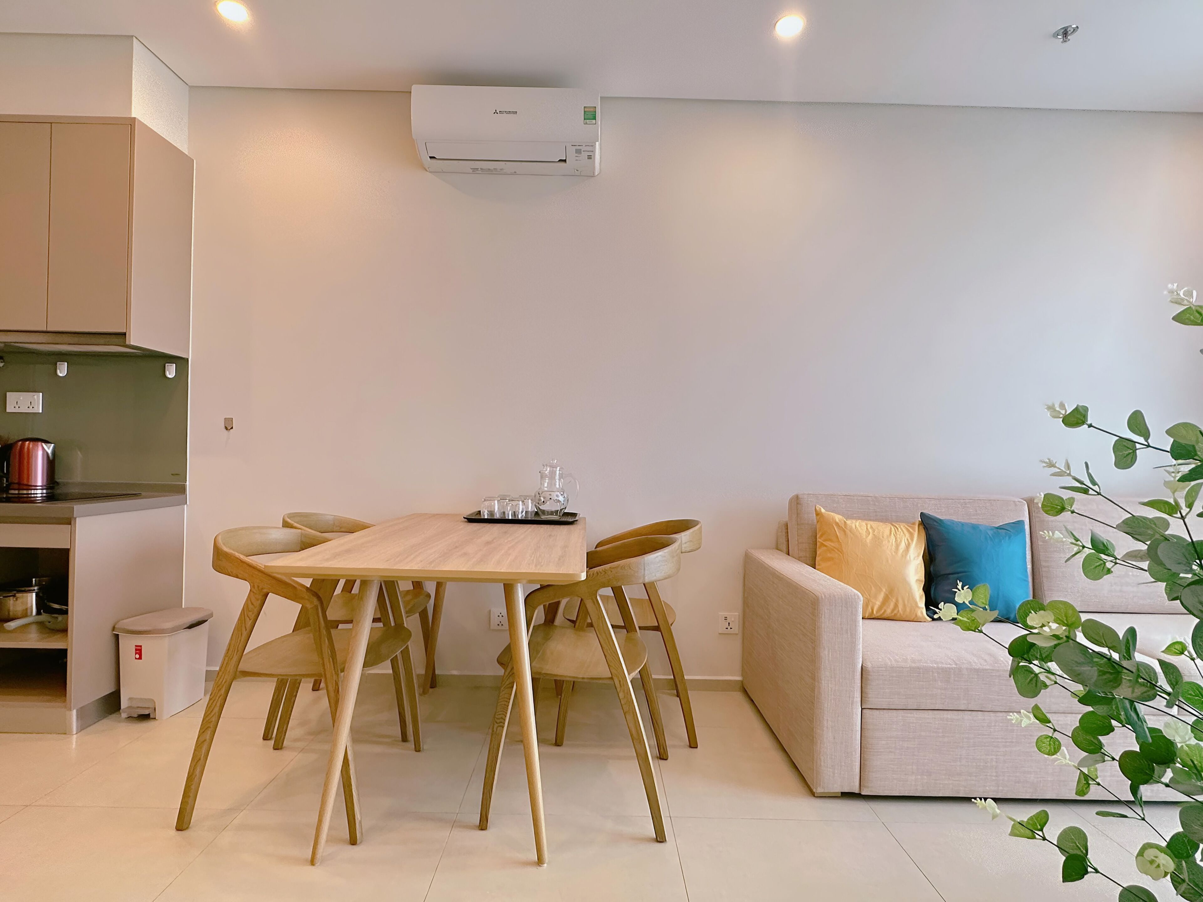 A modern living room with a wooden dining table and chairs, a sofa with cushions, and a kitchen area with a kettle and cabinets.