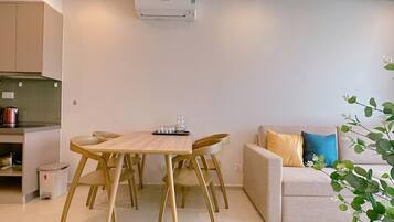 A modern living room with a wooden dining table and chairs, a sofa with cushions, and a kitchen area with a kettle and cabinets.