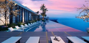 Una piscina infinita moderna con sillones, una terraza de madera y vista al océano al atardecer.