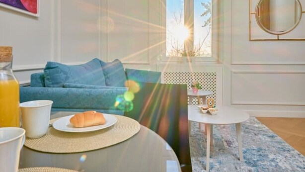 A living room with a blue sofa, a coffee table with a plate of food, a small round table with a plant, and a window with sunlight streaming in. - Salve Luxury Apartment by Litto (Split)