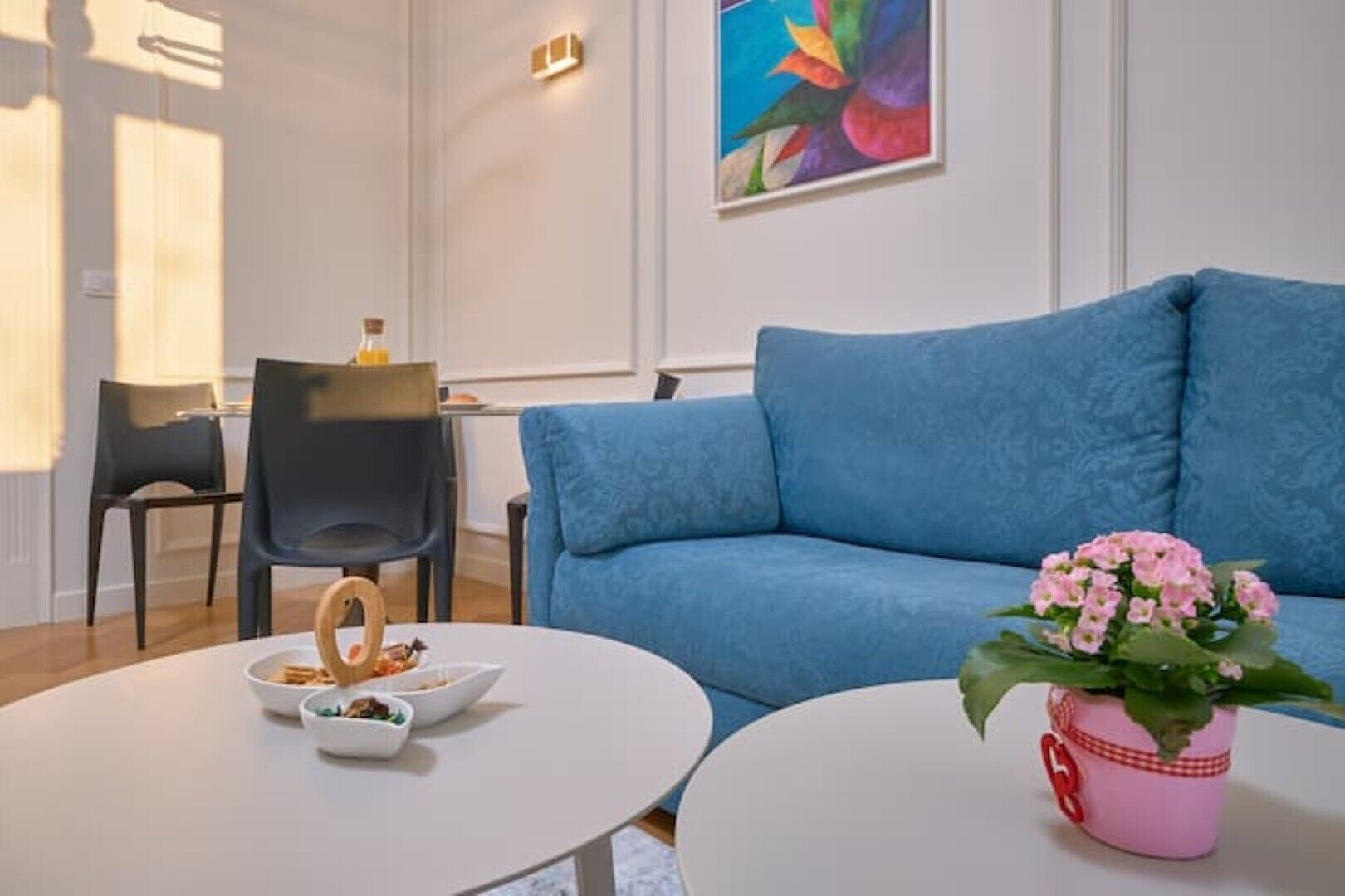 Un salon moderne avec un canapé bleu, une table ronde blanche sur laquelle sont posés des bols, et un pot de fleurs rose.