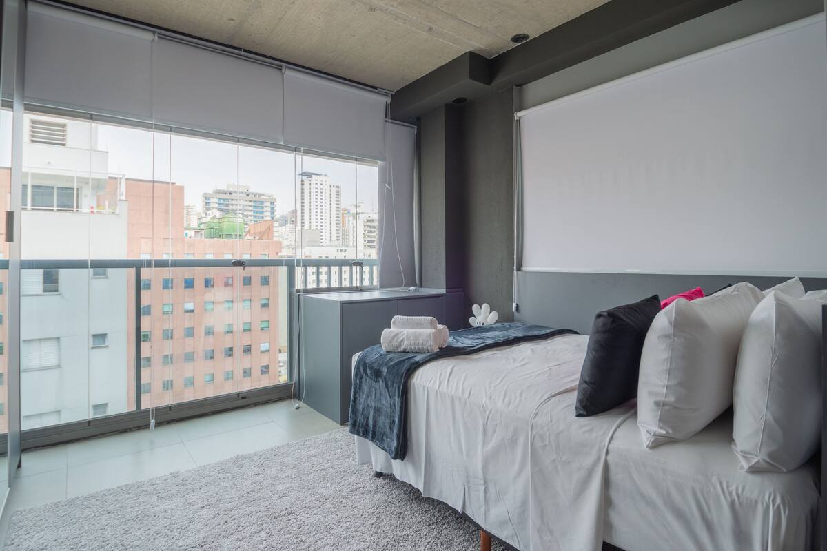 A modern bedroom with a large window, a bed with white bedding, and a view of the cityscape.