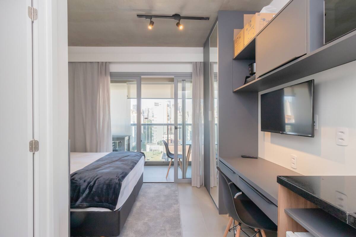 A modern hotel room with a bed, a desk, a TV, and a balcony with a view of the city.