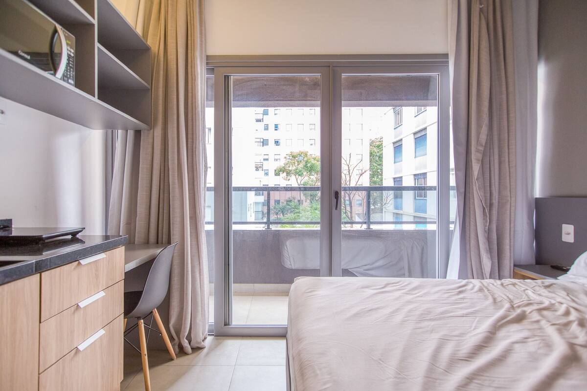 A modern bedroom with a bed, a desk, and a chair. There is a sliding glass door leading to a balcony with a view of buildings and trees.