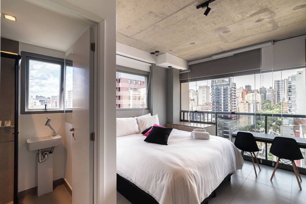 A modern hotel room with a large bed, a small sink, and a balcony with a city view.