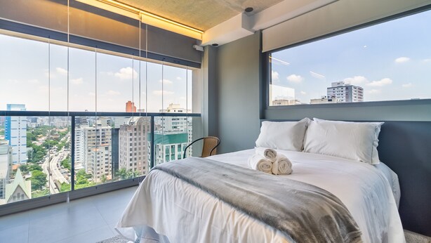 A modern bedroom with a large window, a neatly made bed, and a city view. - 360 VN On Lorena (São Paulo)