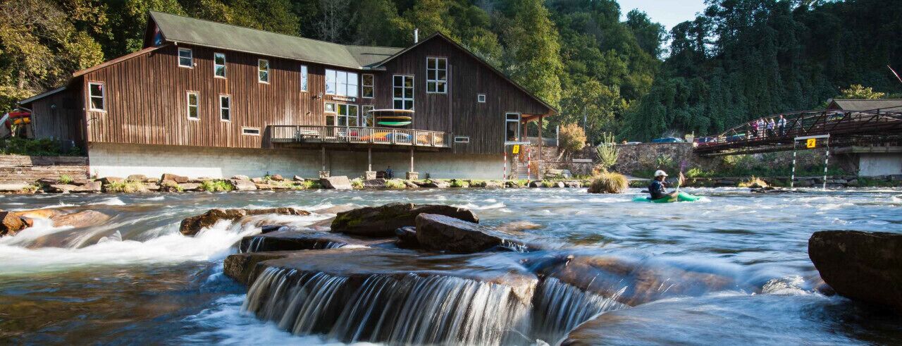 Un bâtiment en bois, avec une rivière en contrebas, une cascade et une personne pratiquant le kayak.