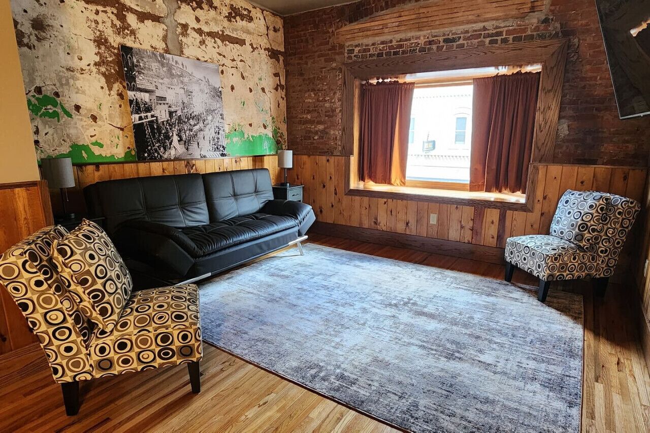 Una stanza con un divano in pelle nera, poltrone a fantasia, pavimento in legno, un'ampia finestra con tende e un muro in mattoni rustici.