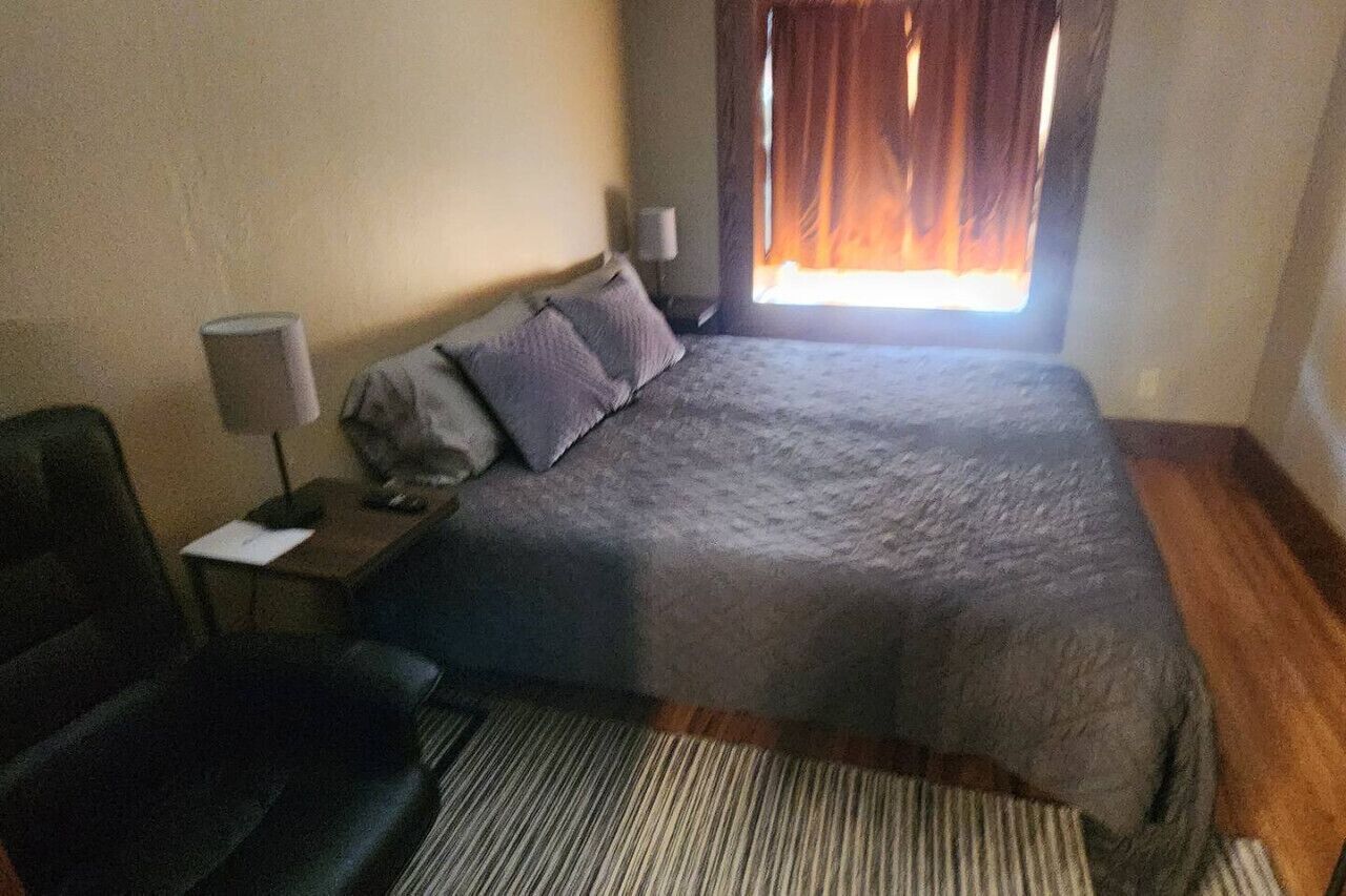 Quarto de hotel com cama, escrivaninha, cadeira, luminária e janela com cortinas.